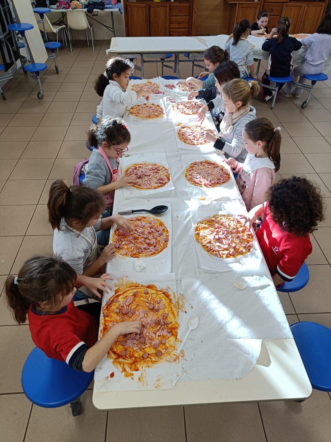 Alumnos de Infantil y Primaria preparando pizzas sobre una mesa larga del comedor
