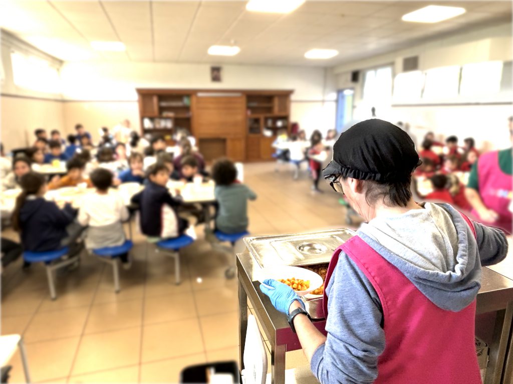 Una empleada del comedor sirve un plato junto al carro caliente, con el comedor lleno al fondo