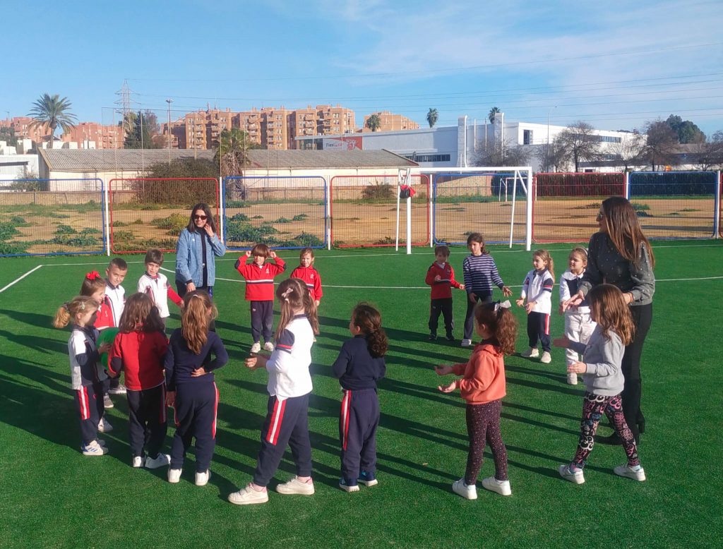 Niños y niñas de Infantil en corro sobre el campo de césped artificial, con sus monitoras