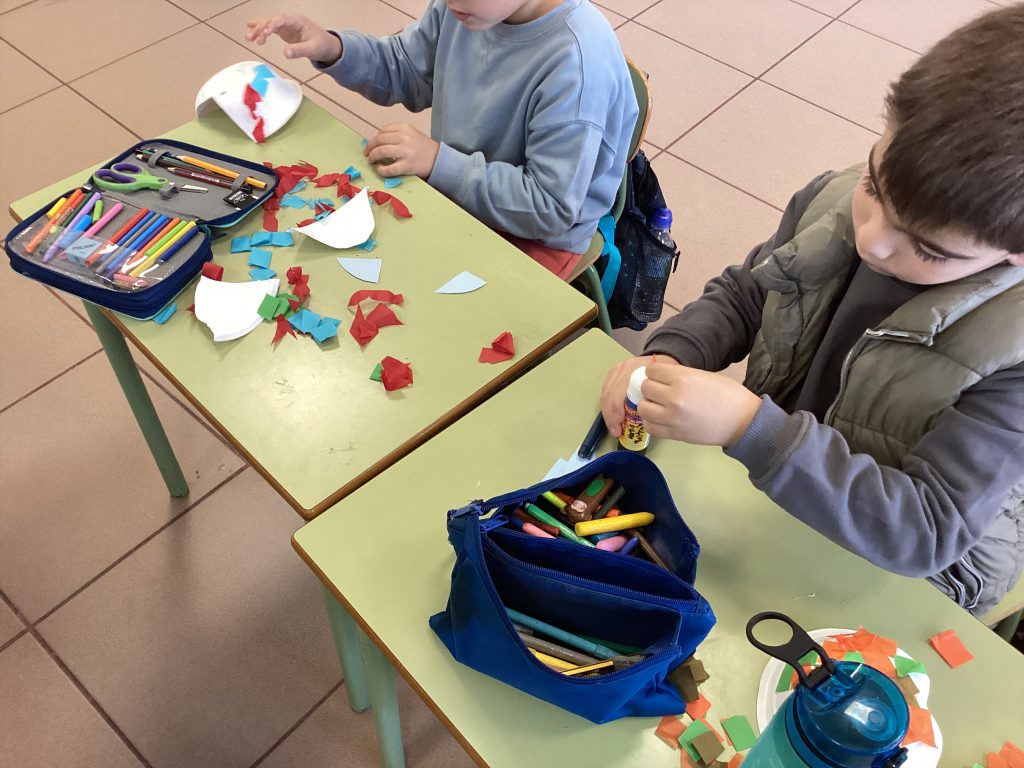 Dos alumnos recortando y pegando papel de colores en las mesas del aula
